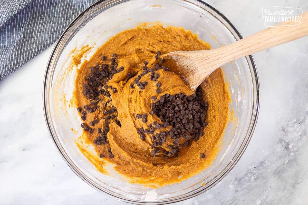 Chocolate chips added to glass mixing bowl of pumpkin chocolate chip muffin batter and a wooden spoon.