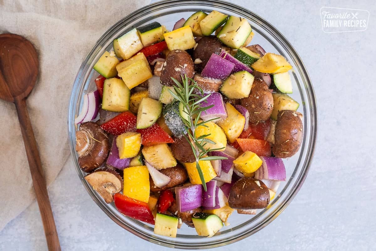 Glass mixing bowl with cut up pieces of zucchini, yellow squash, red onion, red bell pepper and mushroom tossed with seasonings.