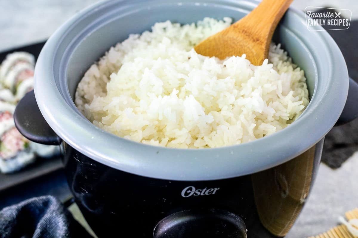 Rice cooker with sushi rice and a wooden spoon with rolled sushi in the background.