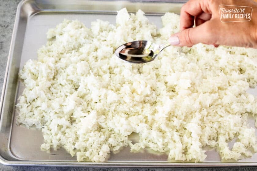 Sprinkling rice vinegar on top of sushi rice spread out on a sheet pan.