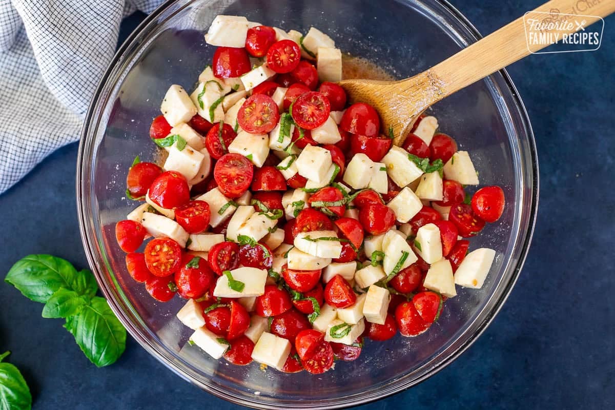 Glass mixing bowl with Tomato Basil Pasta including cherry tomatoes, mozzarella cheese and fresh basil in an olive oil vinegar dressing.