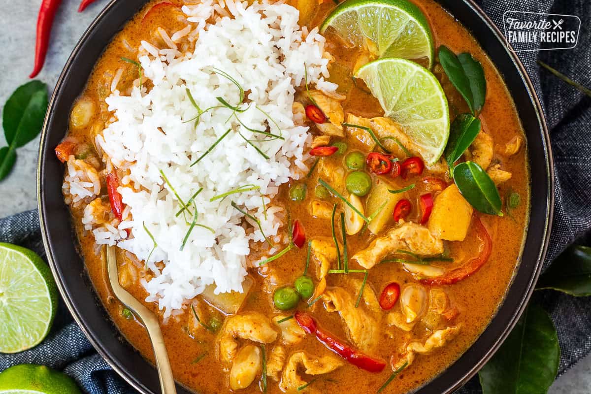 Bowl of chicken panang curry and rice including red bell peppers, peas, potatoes, tomatoes, peas and carrots garnished with lime wedges and Kaffir lime leaves.