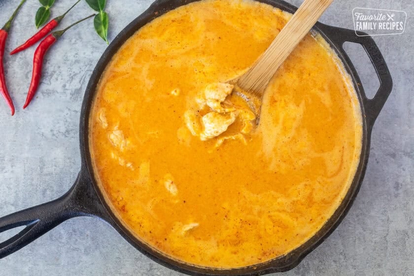 Coconut milk added to panang curry chicken in a skillet with a wooden spoon.