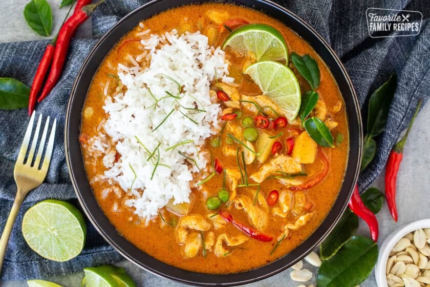 Bowl of chicken panang curry and rice including red bell peppers, peas, potatoes, tomatoes, peas and carrots garnished with lime wedges and Kaffir lime leaves.