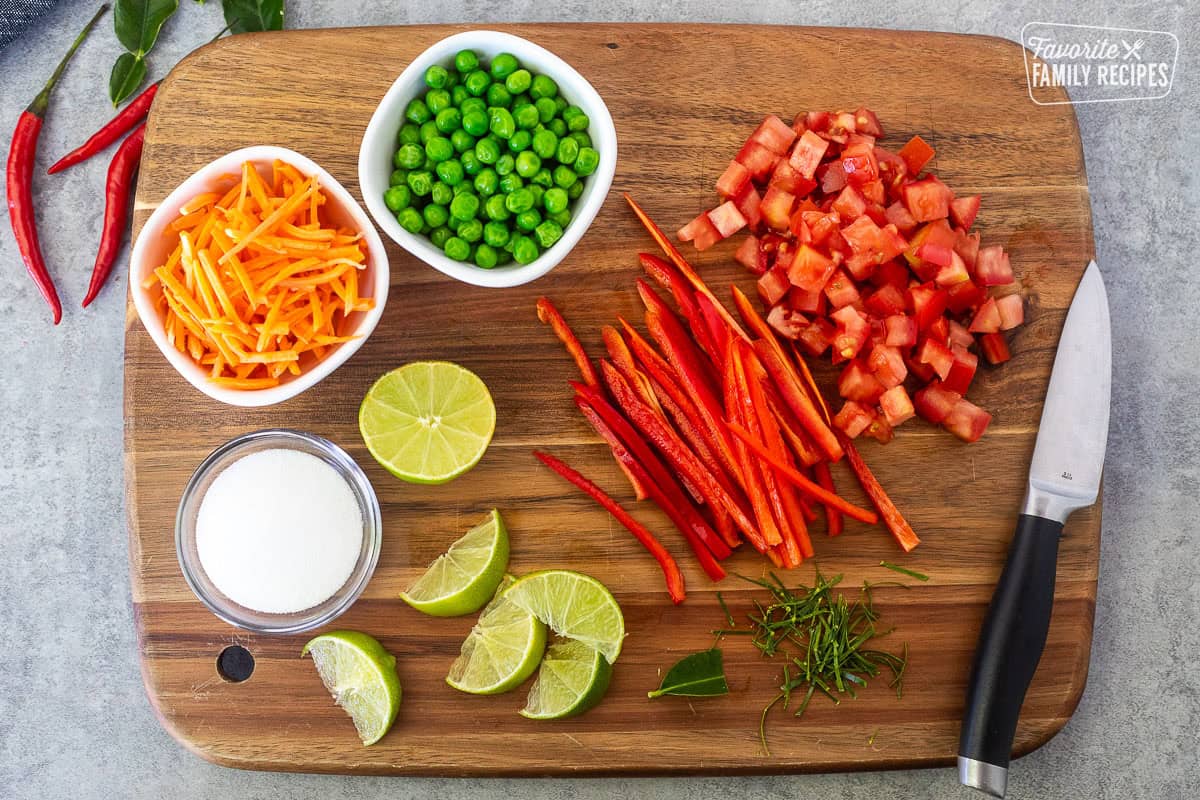 Cutting board with sliced red bell peppers, diced tomatoes, peas, carrots, limes, sugar and Kaffir lime leaves.