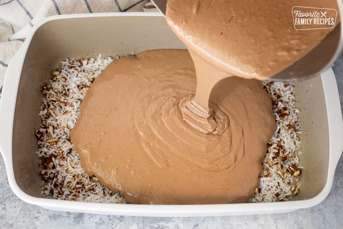 Pouring German chocolate cake mix into baking dish with pecans and coconut flakes.
