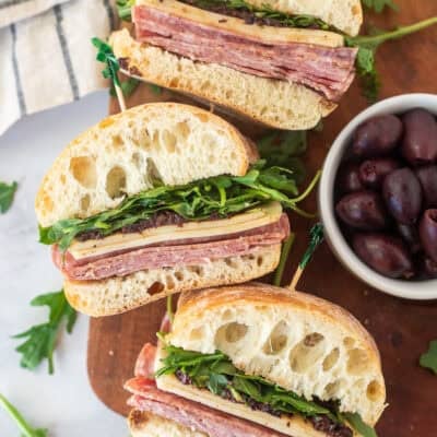 Salami Sandwiches cut in half with salami, provolone cheese, olive tapenade and arugula on a ciabatta roll.
