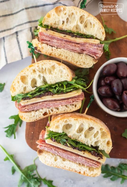 Salami Sandwiches cut in half with salami, provolone cheese, olive tapenade and arugula on a ciabatta roll.