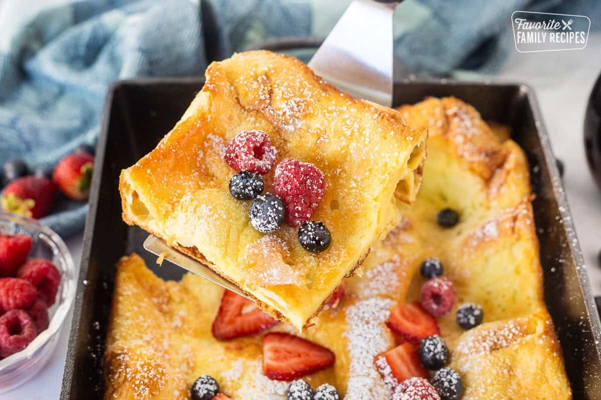 Slice of German pancake on a spatula topped with powdered sugar, sliced strawberries, raspberries and blueberries.