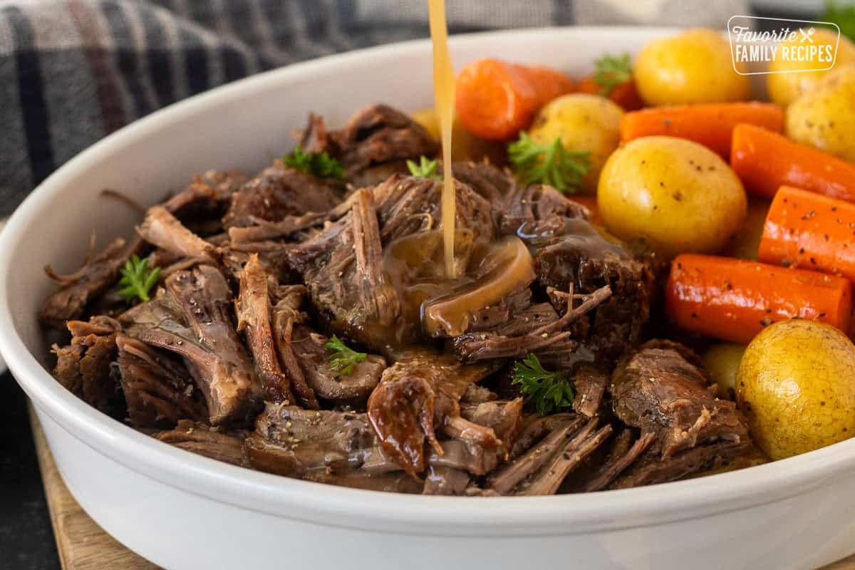 Pouring gravy onto shredded instant pot chuck roast in a dish with cooked vegetables.
