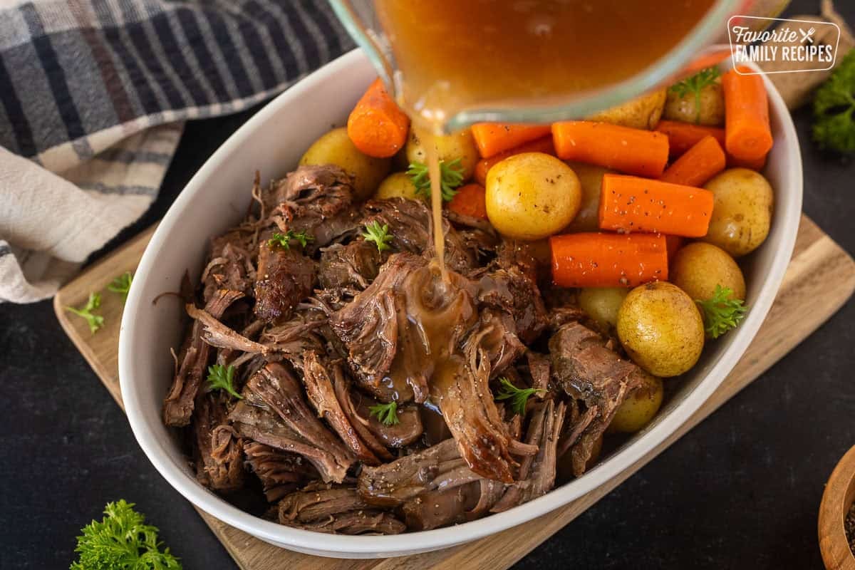 Pouring gravy onto shredded instant pot chuck roast in a dish with cooked vegetables.