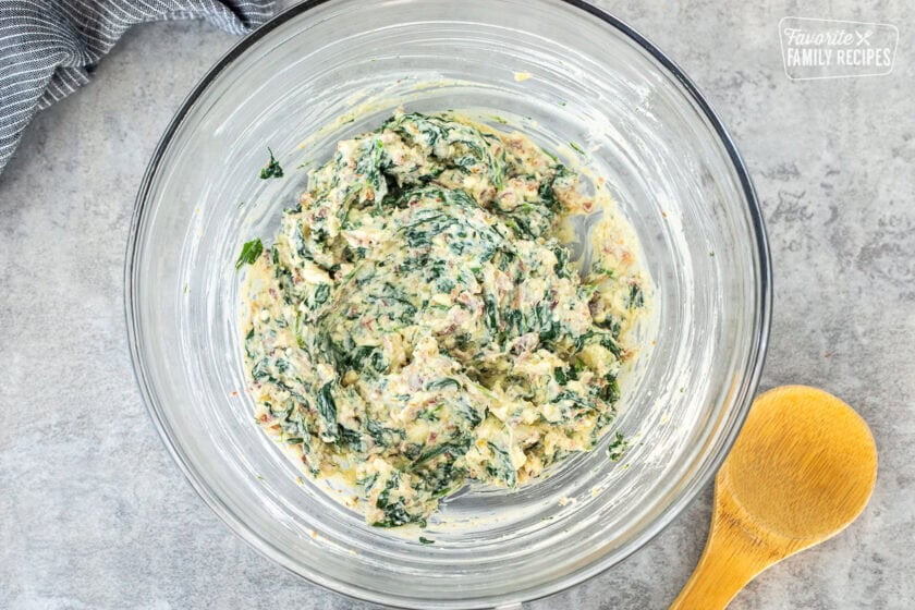 Glass mixing bowl with spinach mixture and wooden spoon on the side.