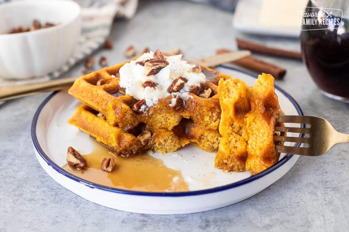 These pumpkin waffles are the perfect fall breakfast. With pumpkin puree, cinnamon, nutmeg, and brown sugar, you get all the warm spices you love packed into a light and fluffy waffle. Two pumpkin waffles cut on a plate with maple syrup, whipped cream and pecans.