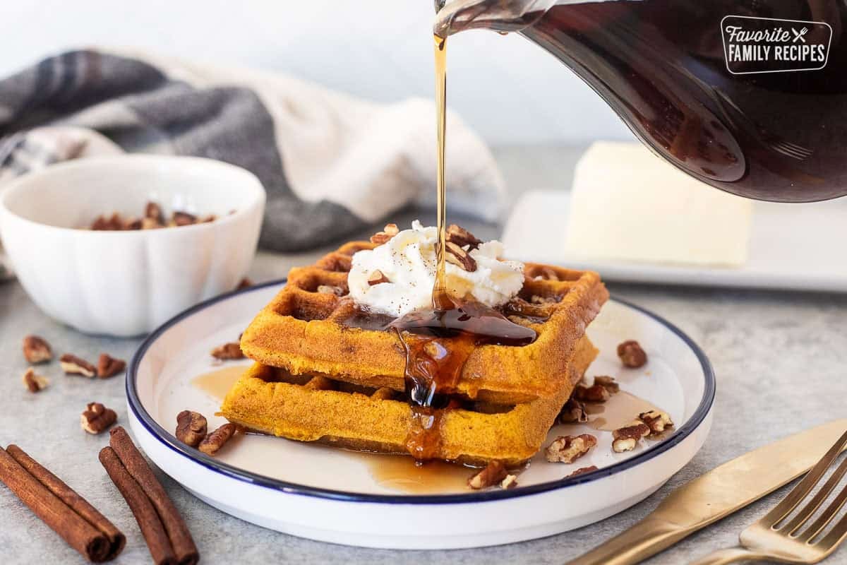 Pouring maple syrup on stacked pumpkin waffles with whipped cream and pecans.