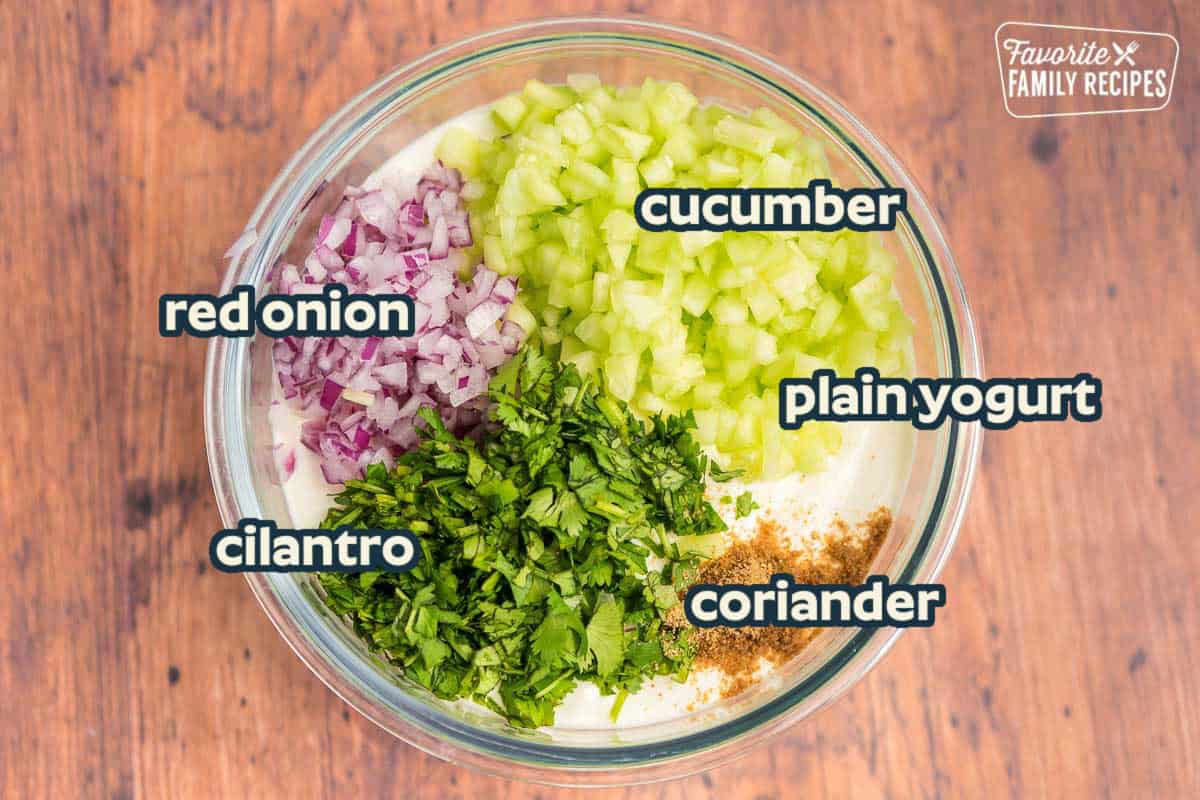 Raita ingredients in a glass bowl: yogurt, cucumber, red onion, cilantro, coriander