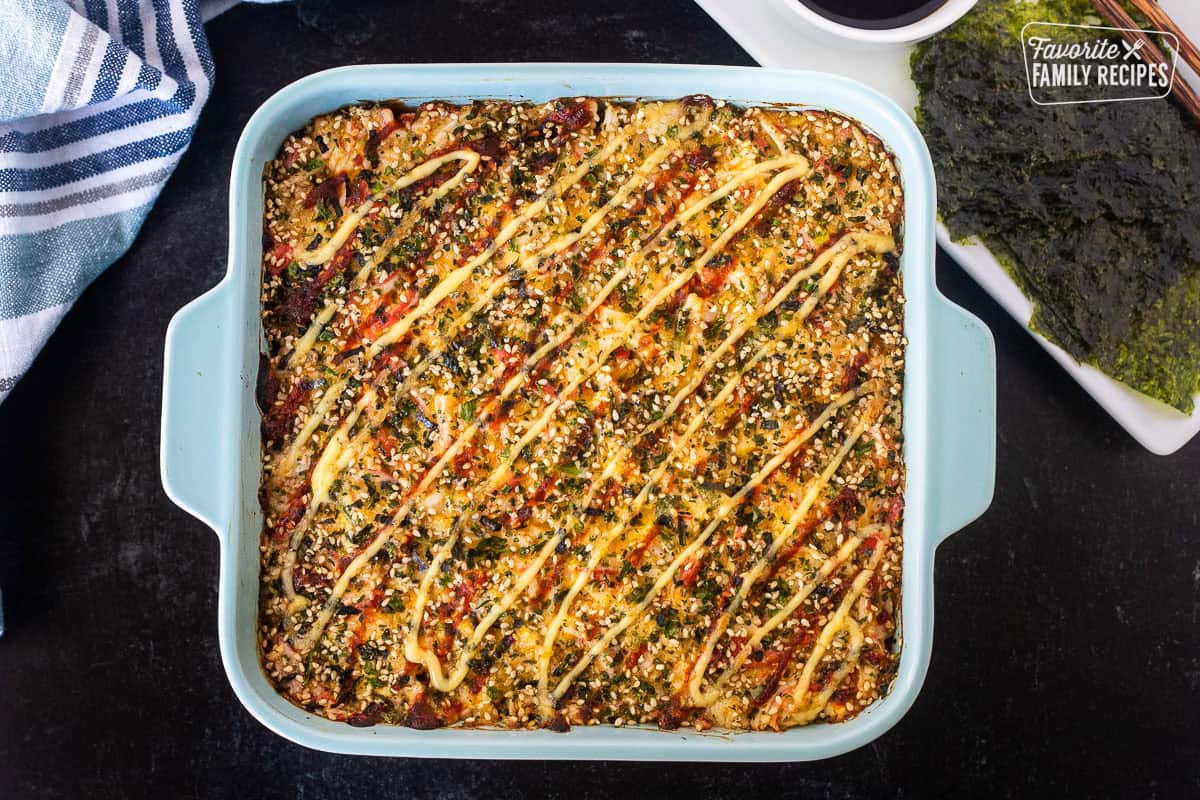 Sushi Bake in a casserole dish with layers of sushi rice, crab, furikake, and spicy mayo with soy sauce and nori on the side.