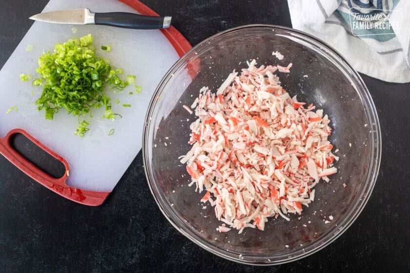 Bowl with shredded imitation crab and sliced green onion on a cutting board to make sushi casserole.