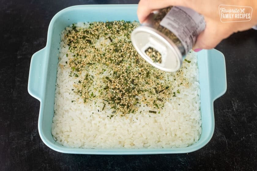 Shaking furikake onto pressed down sushi rice in 8x8 casserole dish.