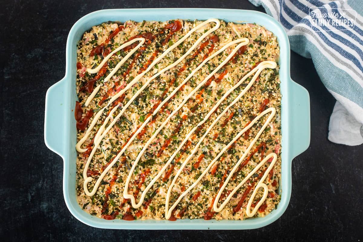 Unbaked sushi bake in a dish topped with furikake, Sriracha sauce and mayo.
