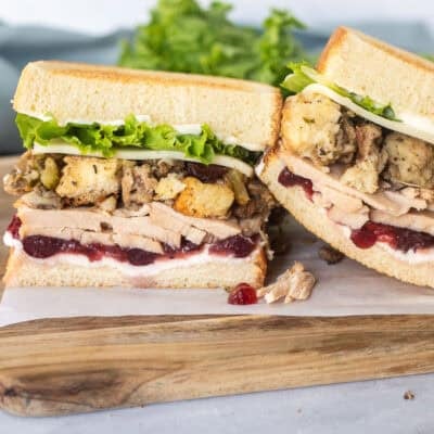 Two halves of Thanksgiving leftover sandwich on toasted white bread with cream cheese, cranberry sauce, sliced turkey, sausage stuffing, provolone cheese, lettuce and mayo.