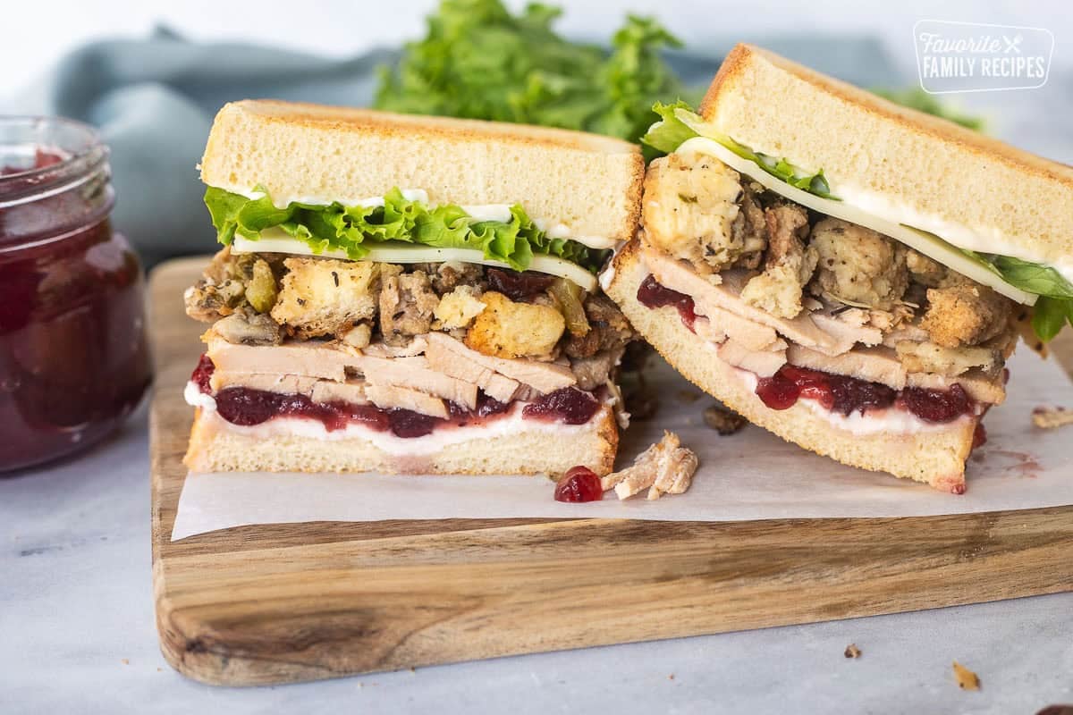 Two halves of Thanksgiving leftover sandwich on toasted white bread with cream cheese, cranberry sauce, sliced turkey, sausage stuffing, provolone cheese, lettuce and mayo.