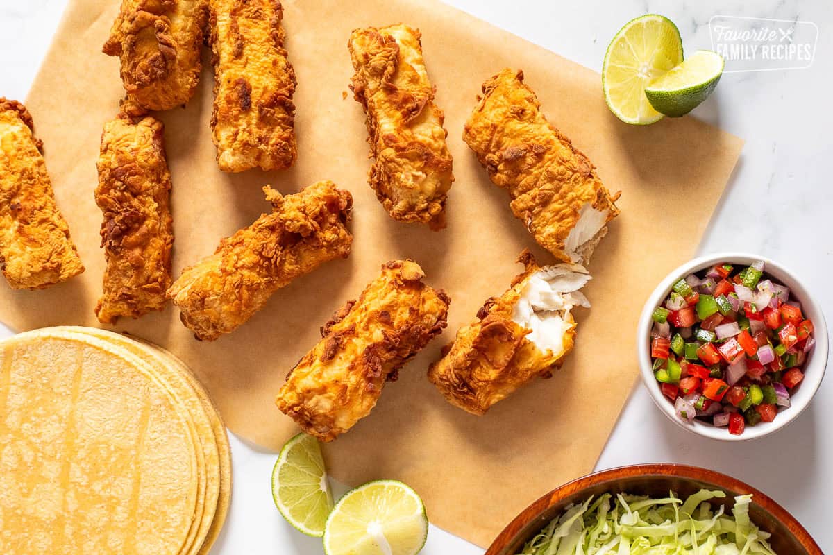 Halibut pieces on parchment paper next to tortillas, salsa, cabbage and limes.