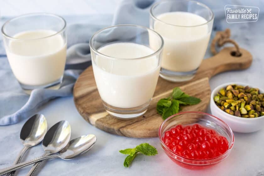 Three glasses of panna cotta with spoons, mint leaves, pistachios and strawberry bursting bubbles on the side.