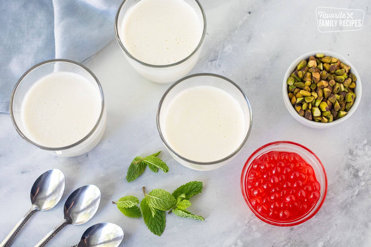 Three glasses of panna cotta with spoons, mint leaves, pistachios and strawberry bursting bubbles on the side.