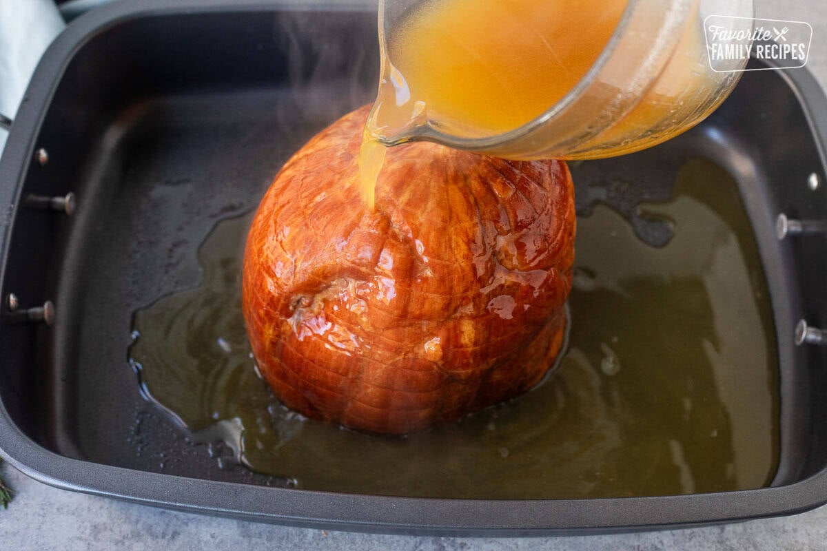Pouring hot apple cider reduction on top of ham in roasting pan.