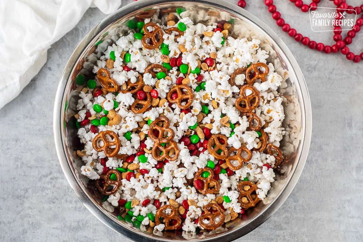 Large bowl of dry Christmas popcorn ingredients combined including popcorn, pretzels, red and green M&M's and roasted peanuts.