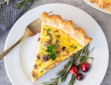 Slice of Christmas Quiche on a plate with rosemary and cranberries on the side.