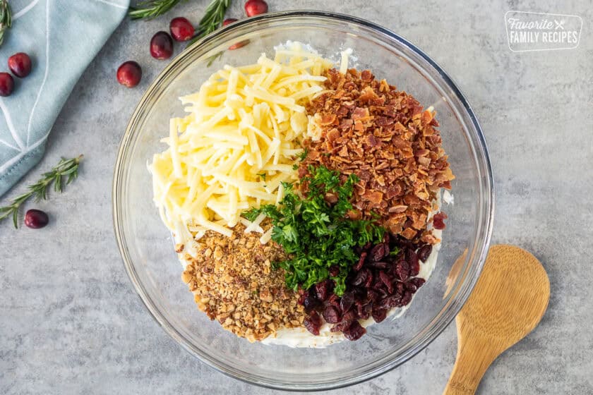 Glass bowl with cream cheese, bacon, dried cranberries, parsley, almonds and shredded sharp white cheddar cheese.