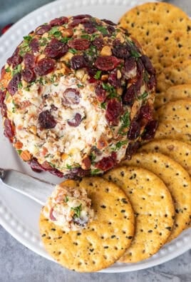 Knife with portion of cranberry cheese ball on top of a cracker next to whole cranberry cheese ball.