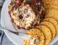 Knife with portion of cranberry cheese ball on top of a cracker next to whole cranberry cheese ball.