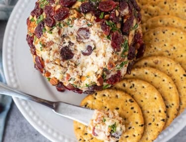 Knife with portion of cranberry cheese ball on top of a cracker next to whole cranberry cheese ball.