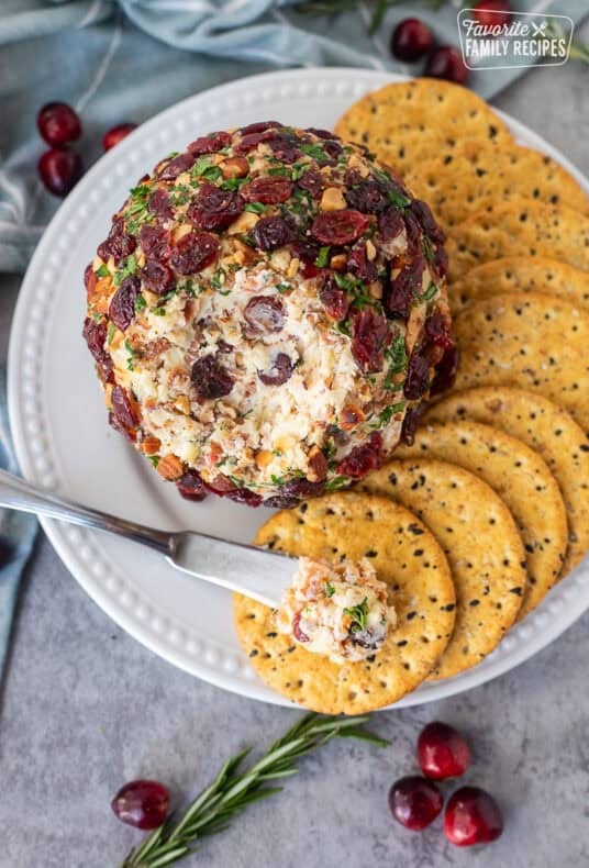 Knife with portion of cranberry cheese ball on top of a cracker next to whole cranberry cheese ball.