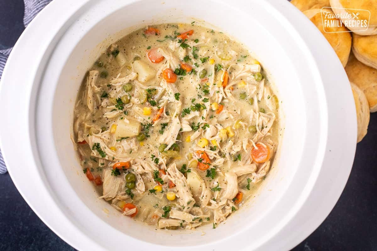 Crock pot chicken pot pie in a crockpot garnished with parsley and pepper with baked biscuits on the side.