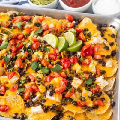 Baked Oven Nachos in a sheet pan topped with tomatoes, green onions, limes and cilantro. Sides of sour cream, salsa and guacamole.
