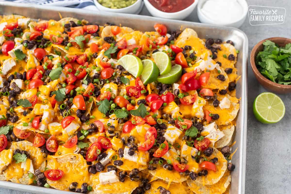 Baked Oven Nachos in a sheet pan topped with tomatoes, green onions, limes and cilantro. Sides of sour cream, salsa and guacamole.