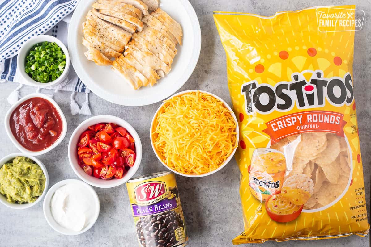 Ingredients to make Oven Nachos including crispy round tortilla chips, shredded cheese, black beans, chicken breast, tomatoes, sour cream, green onions, salsa and guacamole.