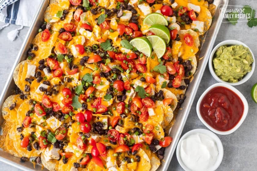 Baked Oven Nachos in a sheet pan topped with tomatoes, green onions, limes and cilantro. Sides of sour cream, salsa and guacamole.