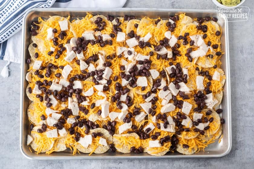 Sheet pan with tortilla chips topped with shredded cheese, chicken pieces and black beans.