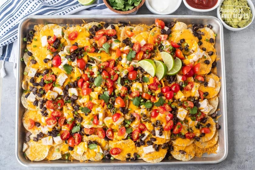 Baked Oven Nachos in a sheet pan topped with tomatoes, green onions, limes and cilantro. Sides of sour cream, salsa and guacamole.