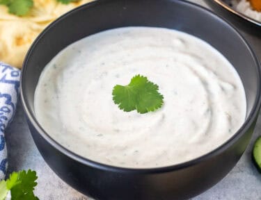 Bowl of Raita sauce next to chicken tikka masala and naan bread. Fresh cucumber and cilantro on the side.