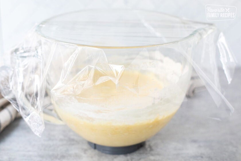 Glass mixing bowl of Sally Lunn Bread dough covered in plastic wrap.