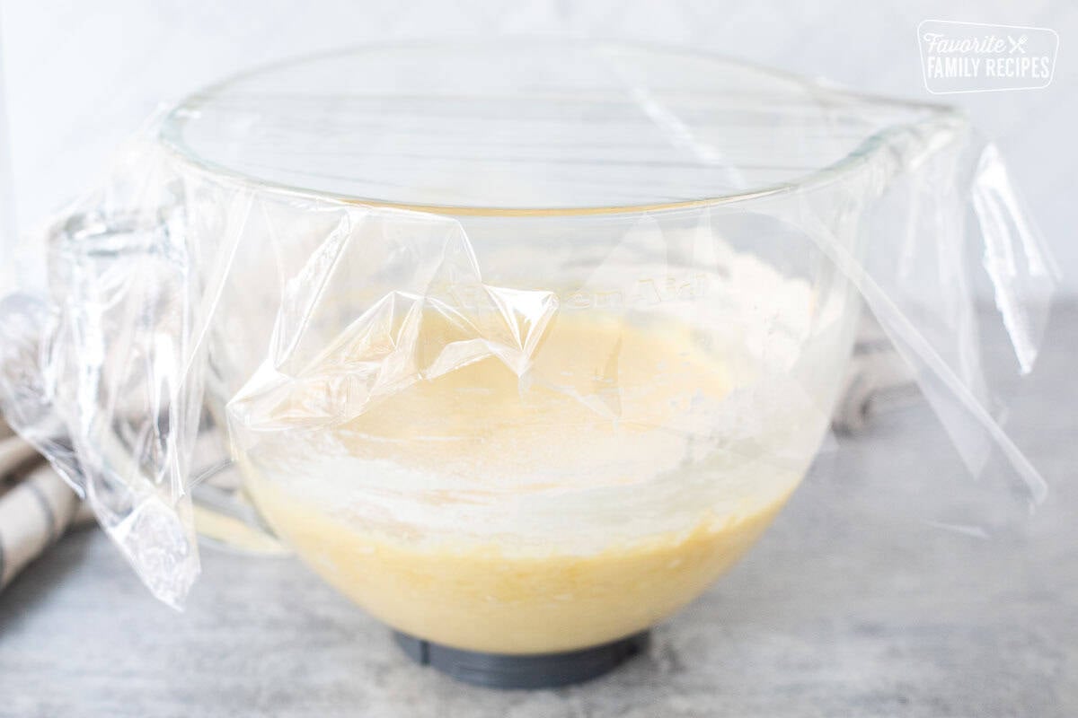 Glass mixing bowl of Sally Lunn Bread dough covered in plastic wrap.