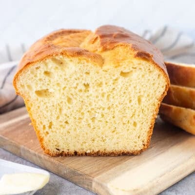 Sliced loaf of Sally Lunn Bread on wooden board. Butter dish and raspberry jam on the side.