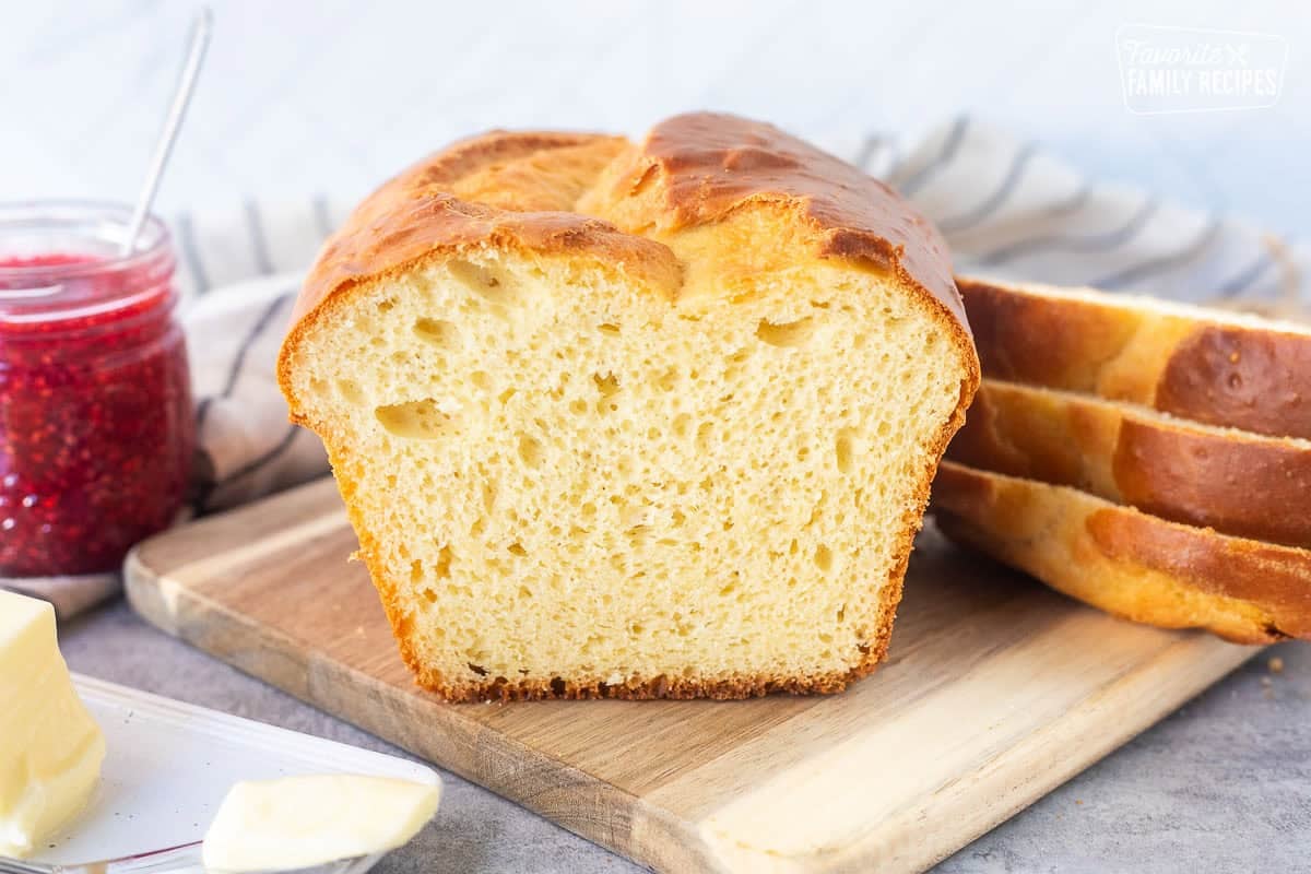 Sliced loaf of Sally Lunn Bread on wooden board. Butter dish and raspberry jam on the side.