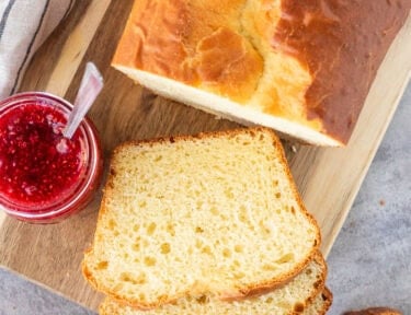 Sally Lunn Bread sliced on wooden board. Butter dish and raspberry jam on the side.