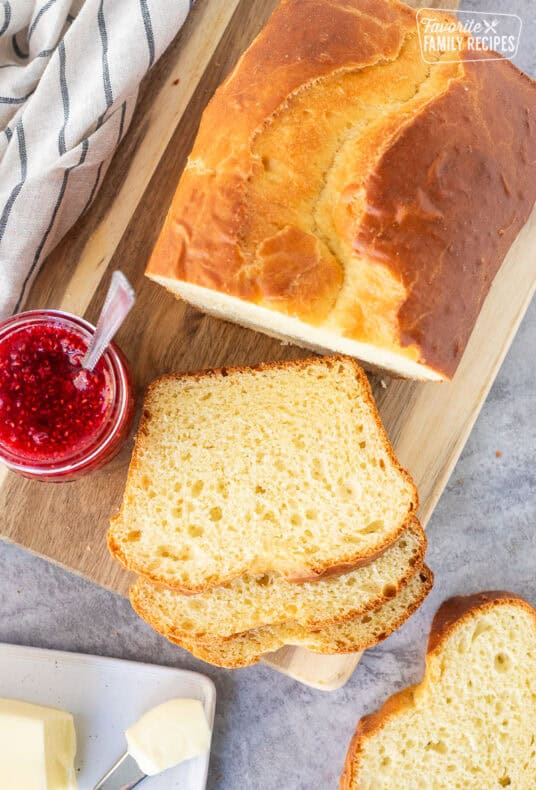 Sally Lunn Bread sliced on wooden board. Butter dish and raspberry jam on the side.